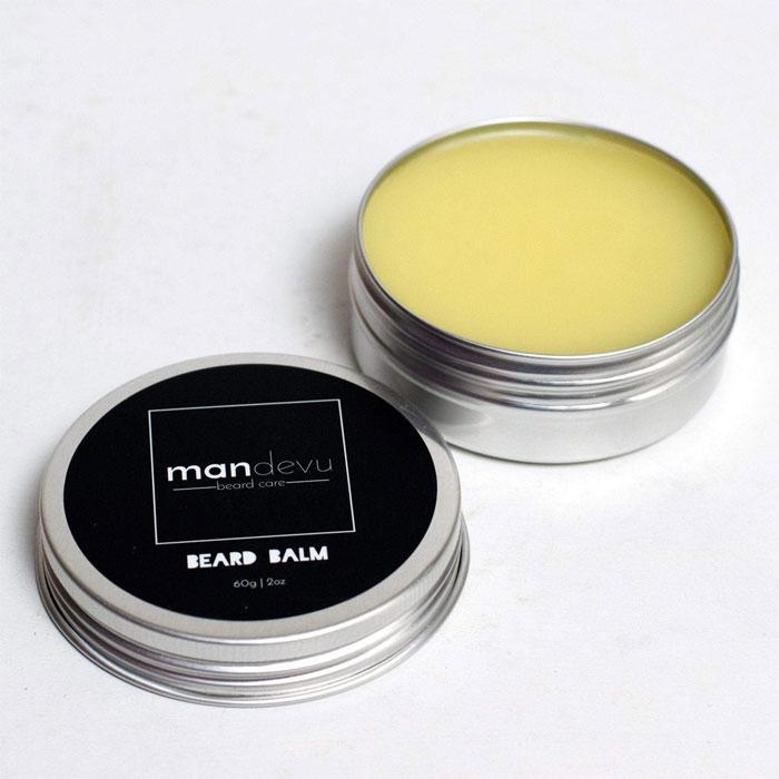 An open metal tin with a black lid, labeled Mandevu Beard Balm, containing a yellow solid substance, presumably the beard balm itself.